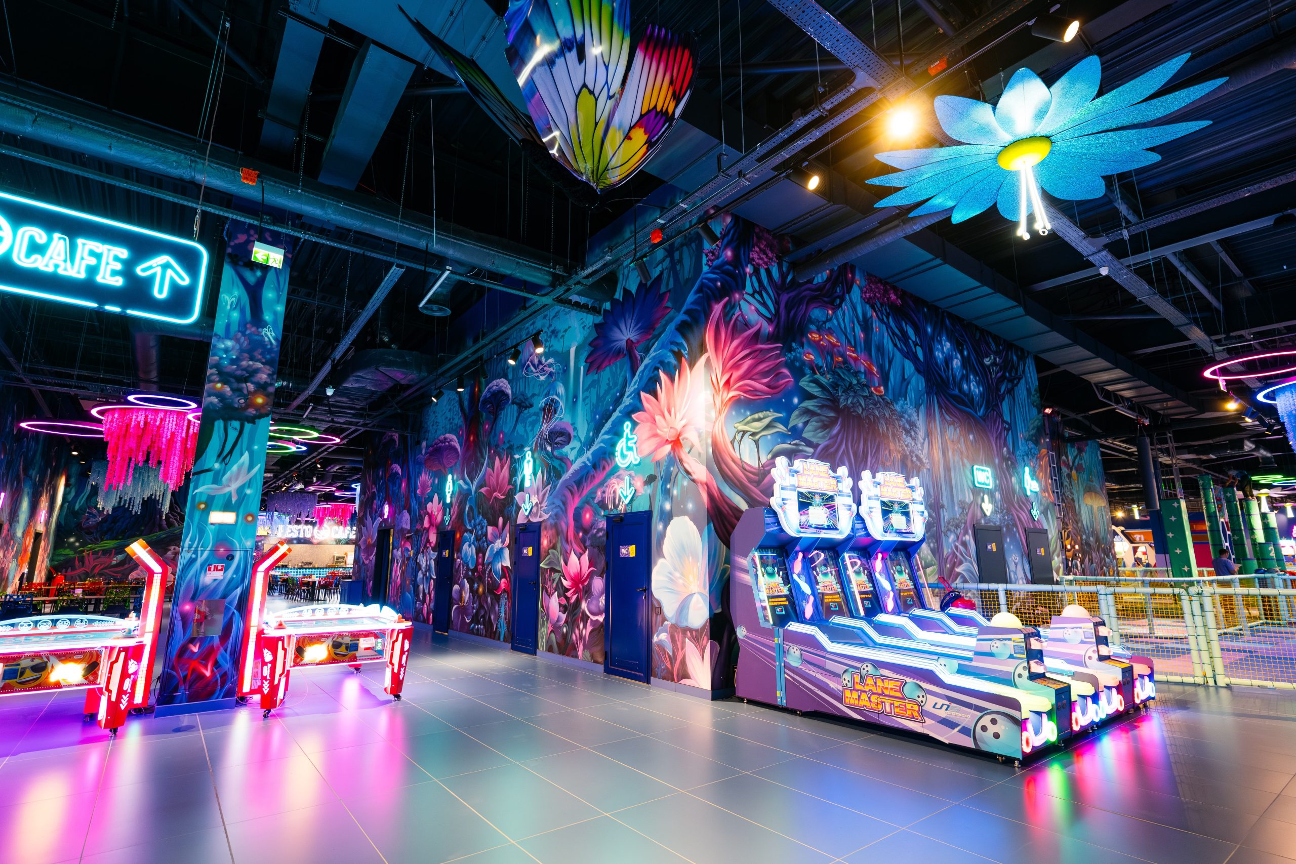 Entertainment venue arcade area with floor-to-ceiling fantasy mural featuring luminescent mushrooms and surreal flora integrated with gaming equipment
