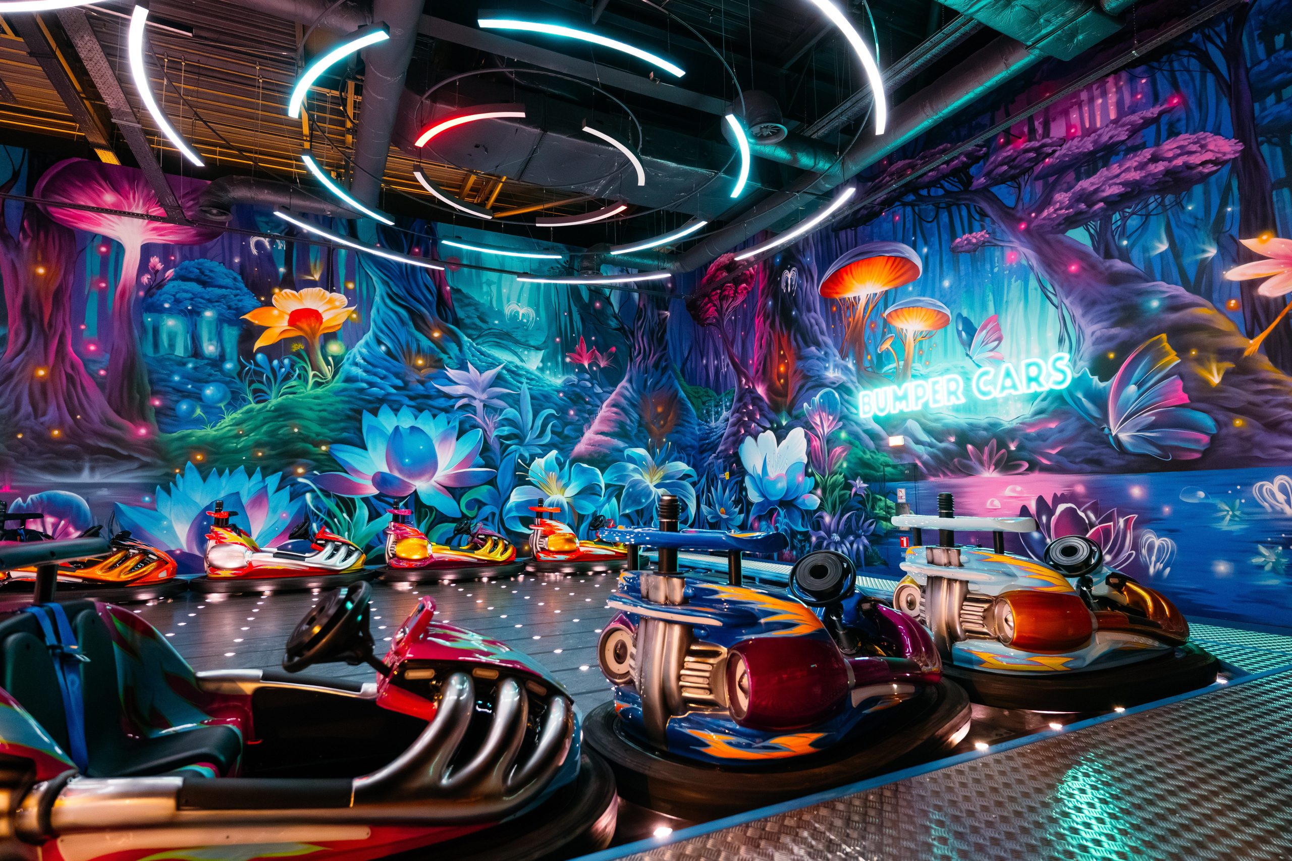 Wide-angle view of bumper car attraction surrounded by 360-degree fantasy mural with neon lighting and bioluminescent flora creating magical atmosphere