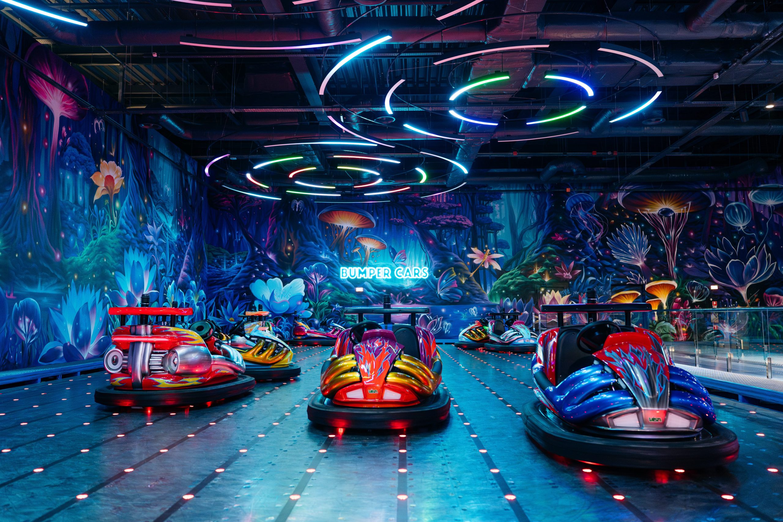 Immersive fantasy wall mural with bioluminescent flora surrounding bumper car area in entertainment venue featuring neon lighting and surreal underwater-themed interior design