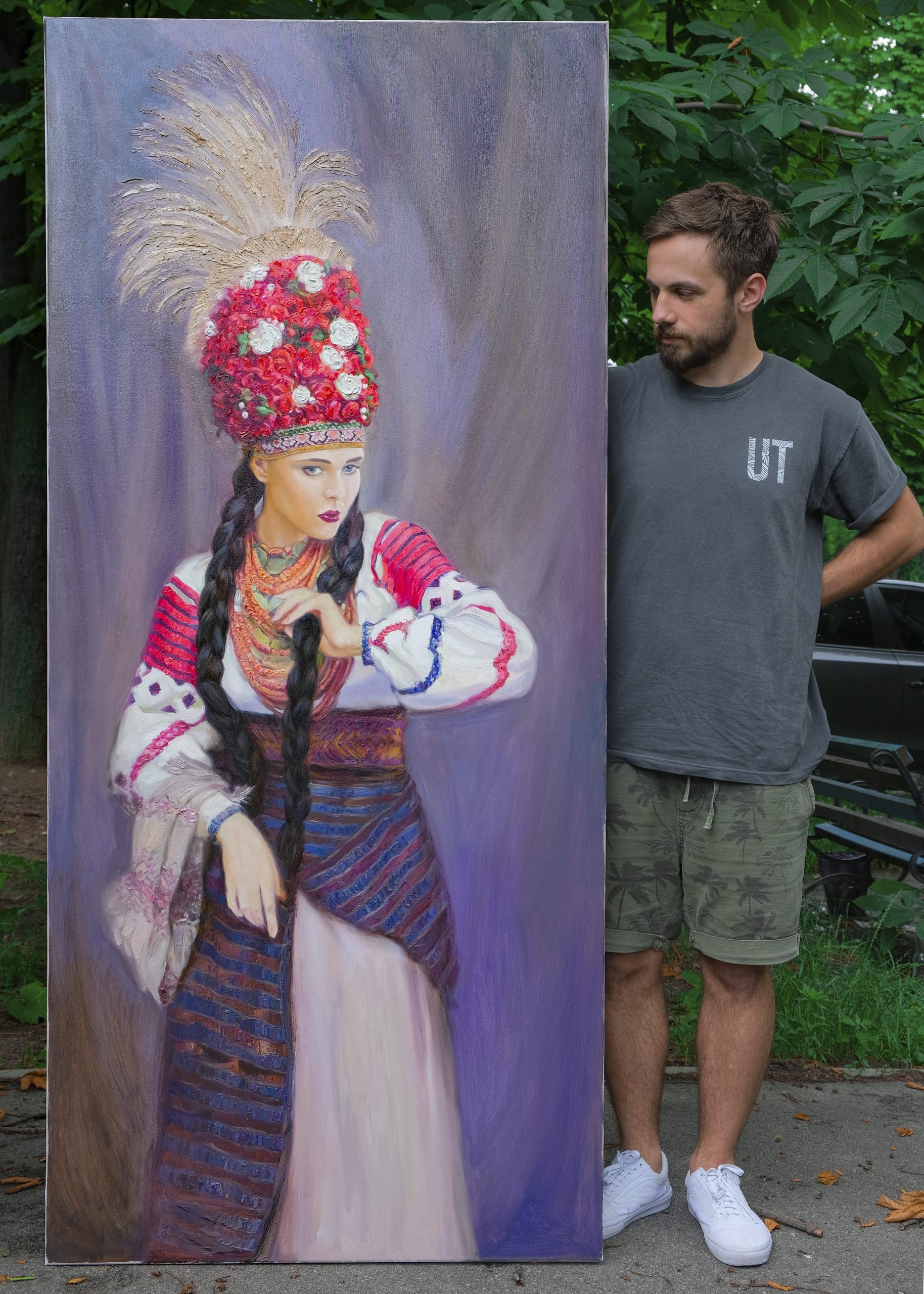 Artist standing next to completed 2-metre life-sized oil portrait on canvas showing full figure of woman in traditional costume with floral crown and layered coral necklaces